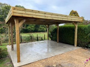 Abri voiture bois, dalle béton, fond vert et ciel nuageux.
